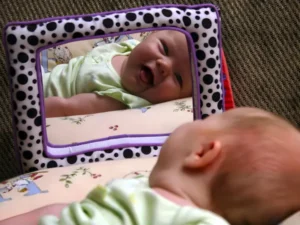 A baby lies on their back, captivated by a toy mirror on the floor