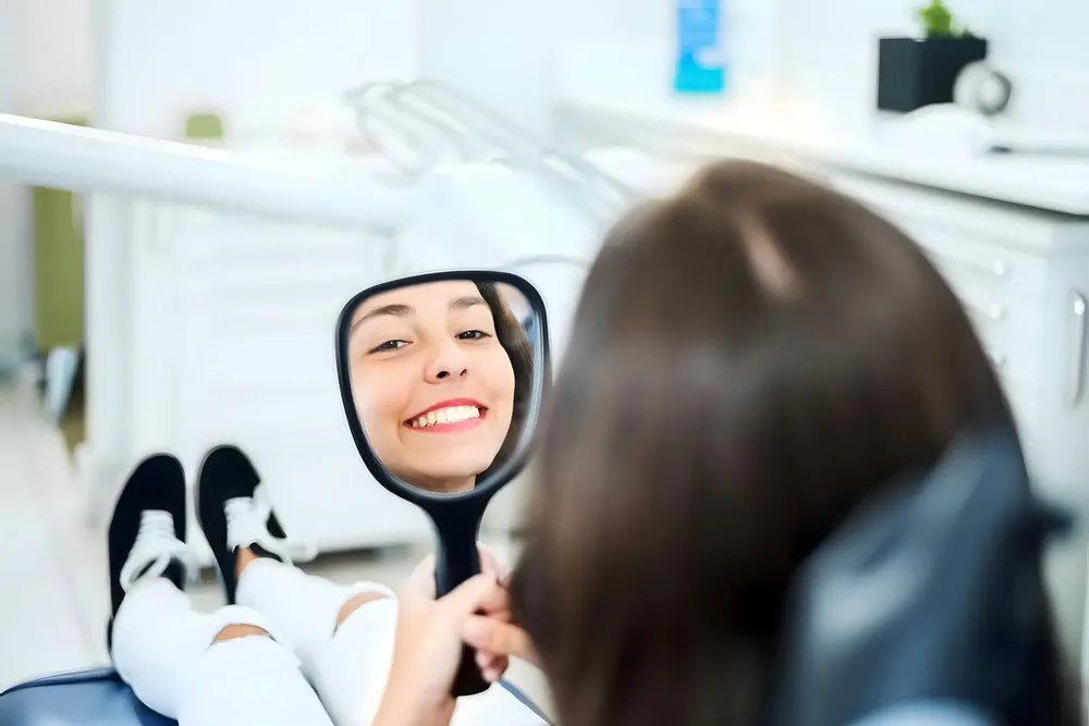 A woman is looking at her teeth with a hand mirror on a chair in a dental clinic