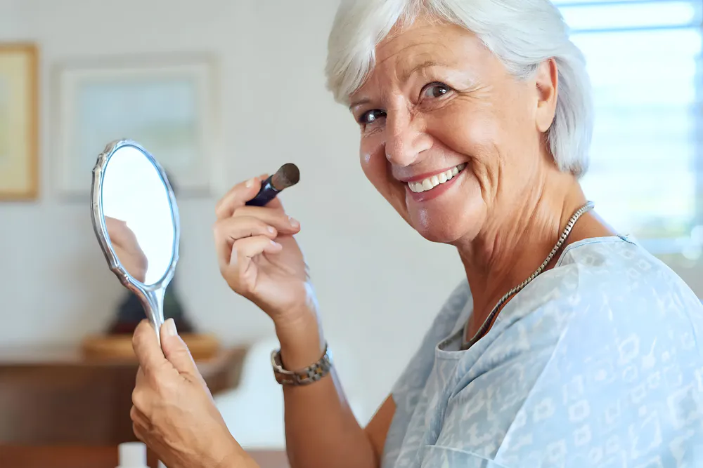 The old lady is touching up her makeup with a hand mirror at home