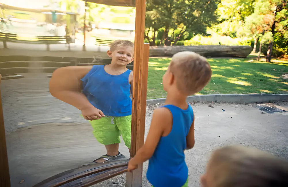 The boy is watching in front of the funhouse mirror
